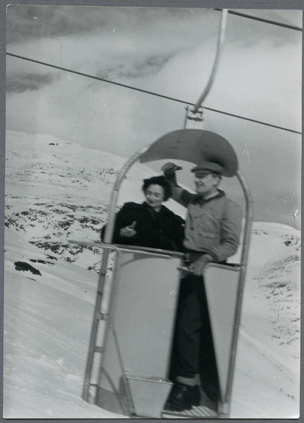 Ein altes Schwarz-Weiß-Foto eines Mannes und einer Frau auf einem Skilift, wobei der Mann eine Mütze trägt und die Frau Schuhe trägt, vor einem schneebedeckten Hintergrund mit sichtbarem Himmel.