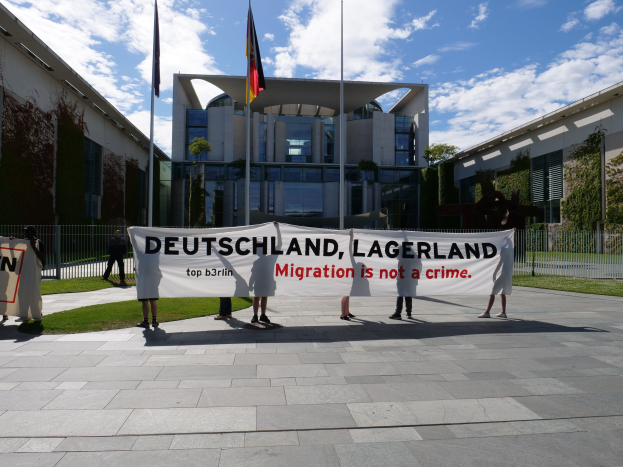 Eine Gruppe von Menschen hält ein Transparent mit der Aufschrift "Deutschland, Lagerland Migration ist kein Verbrechen" vor einem Gebäude mit Metallzaun, umgeben von Gras, Pflanzen, Bäumen und Fahnenmasten, vor einem bewölkten Himmel.
