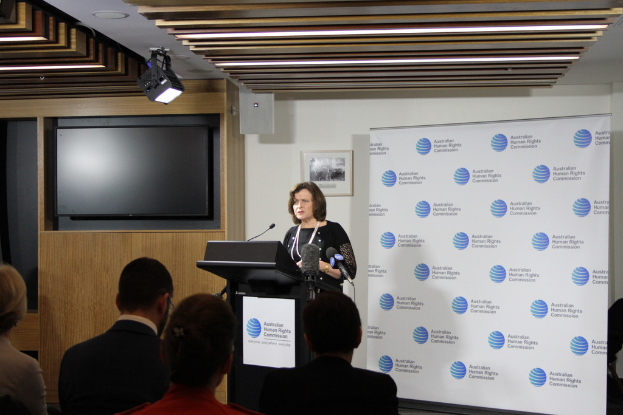 Eine Frau steht auf einer Bühne und spricht in ein Mikrofon, vor ihr sitzen Zuschauer, hinter ihr eine Wand mit einem Banner und einem Fernseher, an der Wand hängen Lampen.