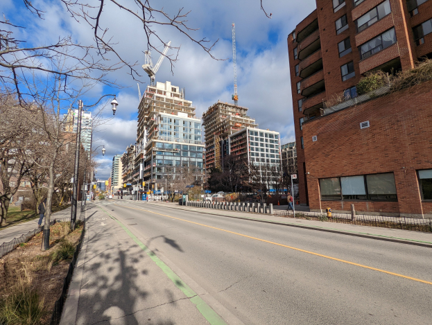 Eine belebte Stadtstraße mit hohen Gebäuden, Straßenlaternen, Stromleitungen, Fahrzeugen, Passanten, Geländern, Bäumen und einem bewölkten Himmel.