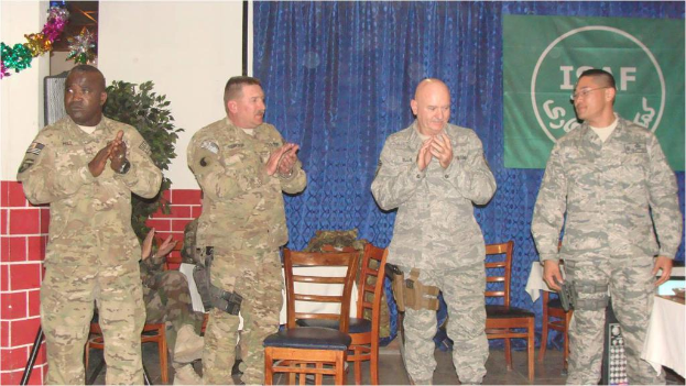 Vier Personen stehen, drei klatschen, einer sitzt auf einem Stuhl neben einer Pflanze, mit einem Banner an einem Vorhang im Hintergrund, alle tragen militärische Kleidung, mit Stühlen und einem Tisch sichtbar.