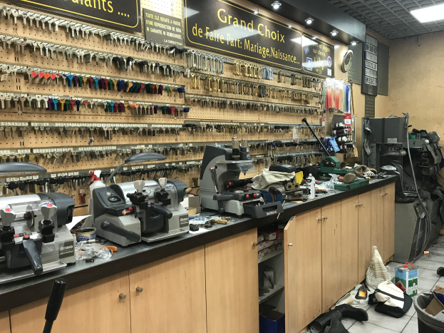 Ein unordentlicher Garage-Workshop mit verschiedenen Werkzeugen, Maschinen, Schränken und Gegenständen auf dem Boden, Tafeln mit Text, eine Wanduhr und Deckenleuchten.