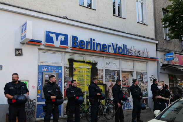 Polizeibeamte vor einer Berliner Volksbank stehend, einige mit Helmen, daneben ein Auto und ein Baum, Gebäude und Texttafeln im Hintergrund.