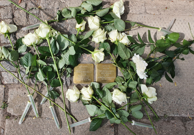 Weiße Rosen und Kerzen in einem Kranz auf dem Boden vor einem Berliner Mahnmal, mit zwei eingravierten goldfarbenen Gegenständen.