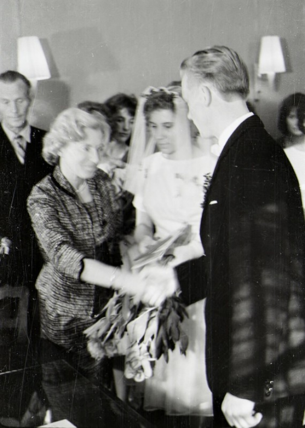 Ein Schwarz-Weiß-Foto einer Braut in einem weißen Kleid und Schleier, die einen Strauß hält, und eines Bräutigams in einem Anzug und Krawatte, die Ringe tauschen, mit Lampen im Hintergrund.