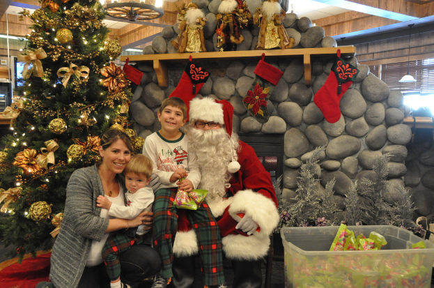 Eine Frau, Kinder und ein Mann in Santa-Klaus-Kostüm mit Brille und Handschuhen stehen neben einem geschmückten Weihnachtsbaum, einer Wand mit Felsen, Spielzeugen, Dekorationen, einem Korb mit Gegenständen und Lichtern im Hintergrund. Ein Junge hält ein Päckchen.