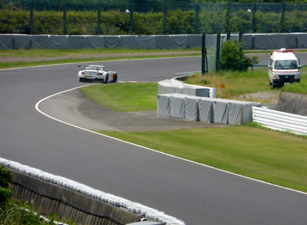 Ein Rennwagen fährt auf einer Rennstrecke, mit einem Polizeiwagen im Hintergrund, umgeben von Gras, Pflanzen, Bäumen und Absperrungen, mit einem Fahrzeug auf der rechten Seite und Text unten.