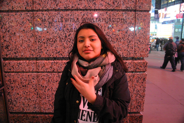 Eine Frau in schwarzer Jacke und Schal steht vor einer Wand, mit vielen Menschen, die auf einem Fußweg rechts an ihr vorbeigehen, und Gebäuden im Hintergrund.