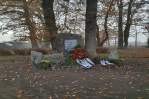 Denkmal in einem Park mit verstreuten trockenen Blättern, einer Texttafel, Blumensträußen und anderen Gegenständen auf Steinen, unter einem sichtbaren Himmel.