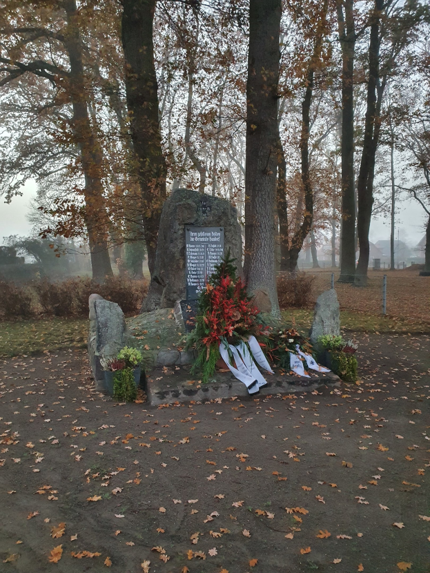Denkmal in einem Park mit verstreuten trockenen Blättern, einer Texttafel, Blumensträußen und anderen Gegenständen auf Steinen, unter einem sichtbaren Himmel.