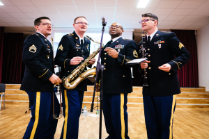 Vier Männer in Militäruniformen stehen zusammen mit Saxophonen, Mikrofon und Flasche in einer bühnenähnlichen Umgebung mit Stühlen, Vorhängen und Deckenlampen.