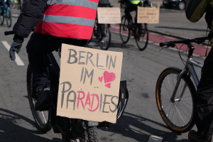 Gruppe von Menschen, die auf Fahrrädern die Straße entlangfahren, mit einem "Berlin I'm Paradies"-Schild im Vordergrund und einem Auto im Hintergrund, Bild etwas unscharf.