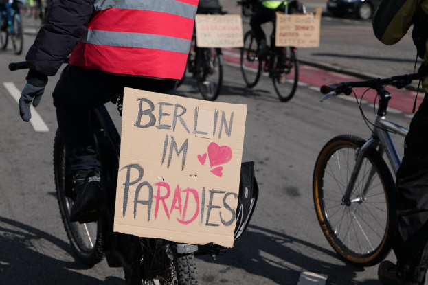 Gruppe von Menschen, die auf Fahrrädern die Straße entlangfahren, mit einem "Berlin I'm Paradies"-Schild im Vordergrund und einem Auto im Hintergrund, Bild etwas unscharf.