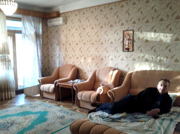 Ein Wohnzimmer mit einem Kronleuchter, Vorhängen, einer Tür, einer Klimaanlage, einem Rahmen an der Wand, einer Couch mit einer Person darauf, einem Teppich und einem Tisch.
