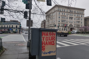Eine belebte Stadtstraße mit Fahrzeugen, einem Fußgänger, einem Müllcontainer mit einer 'Waschen Sie Ihre Hände'-Aufschrift, Verkehrsampeln, Schildern, einem Metallzaun, Pflanzen, Bäumen, Gebäuden und einem klaren blauen Himmel.