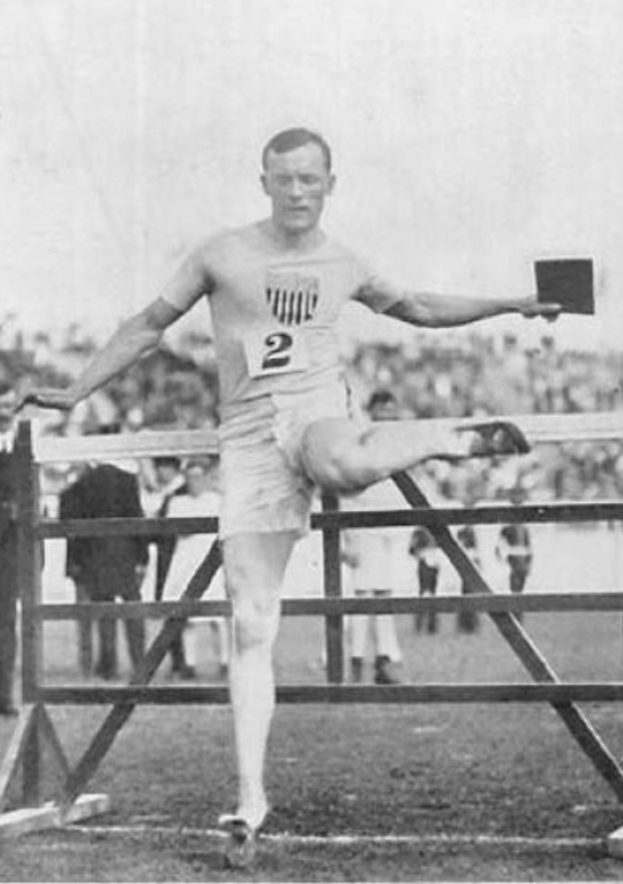 Schwarz-weiß-Foto eines Mannes in Sportkleidung, der über ein Hindernis springt, mit einer Nummer auf seinem Hemd und Shorts, vor einem Publikum mit dem Himmel im Hintergrund.