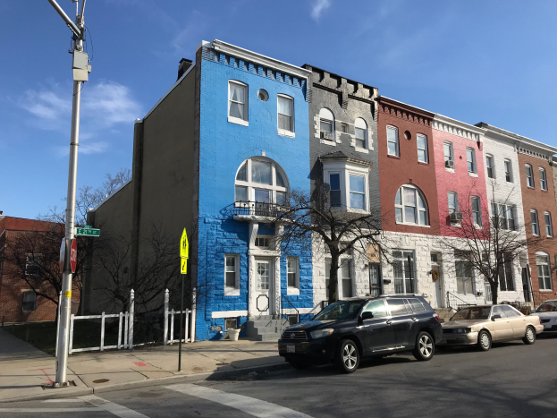 Eine Reihe bunter Häuser mit parkenden Autos auf einer Straße, die von Bäumen, Pfählen, Brettern und einem Zaun gesäumt ist, unter einem bewölkten Himmel.