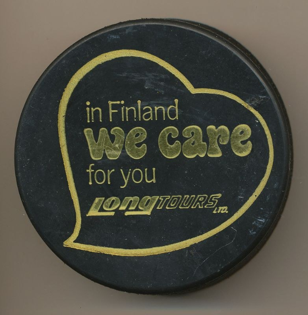 Schwarzer Hockey-Puck mit der Aufschrift "In Finland We Care For You Long Tours" auf einem cremefarbenen Hintergrund.