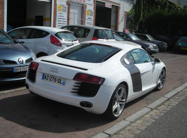 Ein weißer Audi R8 steht auf einer Straße vor einem Gebäude mit vernagelten Fenstern, umgeben von Bäumen auf der rechten Seite.