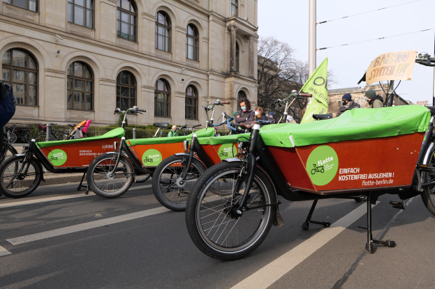 Gruppe von Fahrrädern, die auf der Seite einer Straße geparkt sind, mit einer Person, die links steht, Gebäuden und Bäumen im Hintergrund unter einem klaren blauen Himmel, Teil einer Fahrrad-Sharing-Kampagne, wie durch ein Banner angezeigt.