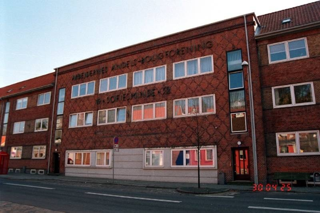 Ein großes Ziegelgebäude mit vielen Fenstern, identifiziert als Hauptquartier der deutschen Bundespolizei, das sich auf einer Straße mit einem Gehweg, einem Baum, einer Bank, einem Laternenmast und einem roten Kasten befindet, mit dem Himmel im Hintergrund.