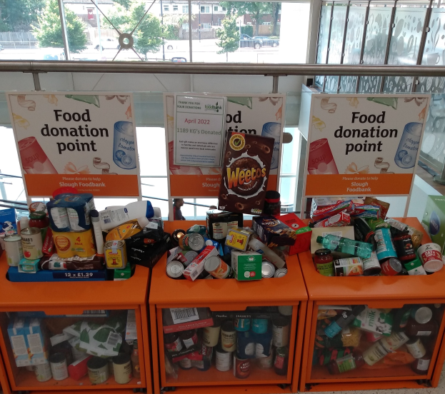 Orange Schränke voller gespendeter Lebensmittel wie Flaschen, Dosen und Kartons, mit Texttafeln daneben, durch Glasfenster betrachtet, die Fahrzeuge, Bäume, Gebäude und Himmel zeigen.