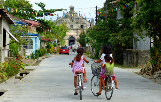Drei Kinder fahren Fahrräder auf einer von Bäumen gesäumten Straße mit Gebäuden auf beiden Seiten, im Hintergrund ein Auto und ein Gebäude und oben der Himmel