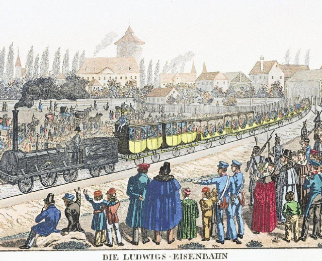 Eine Gruppe von Menschen steht in der Nähe eines Zuges auf den Schienen, einige sitzen im Zug und andere auf dem Boden, vor Gebäuden und Bäumen, mit dem Text "Ludwigs-Eisenbahn, der erste Bahnhof in Deutschland" unten.