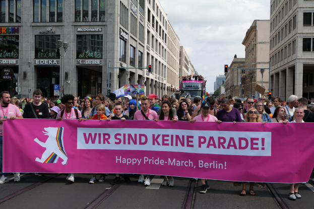 Eine Gruppe von Menschen marschiert auf einer Straße in Berlin, Deutschland, mit einem pinken "Happy Pride March"-Schild, während Gebäude, Laternenpfähle und Verkehrszeichen die Straße säumen, unter einem bewölkten Himmel.