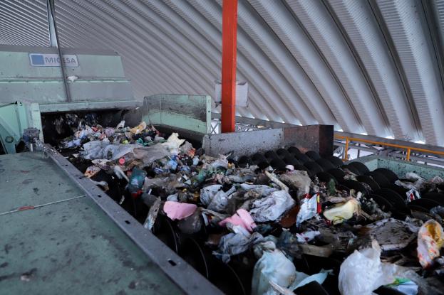 Ein großer Müllberg auf einem Förderband in einer Recyclinganlage, mit Maschinen im Vordergrund und einem Metalldach darüber.