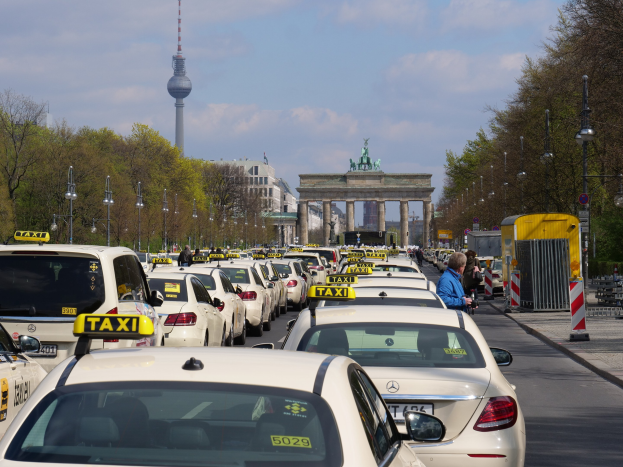 Eine belebte Straße in Berlin mit vielen geparkten Taxis, Fußgängern auf dem Gehweg, Laternenpfählen, Bäumen, Gebäuden, einem Bogen mit Statuen, einem Turm in der Ferne und einem bewölkten Himmel.