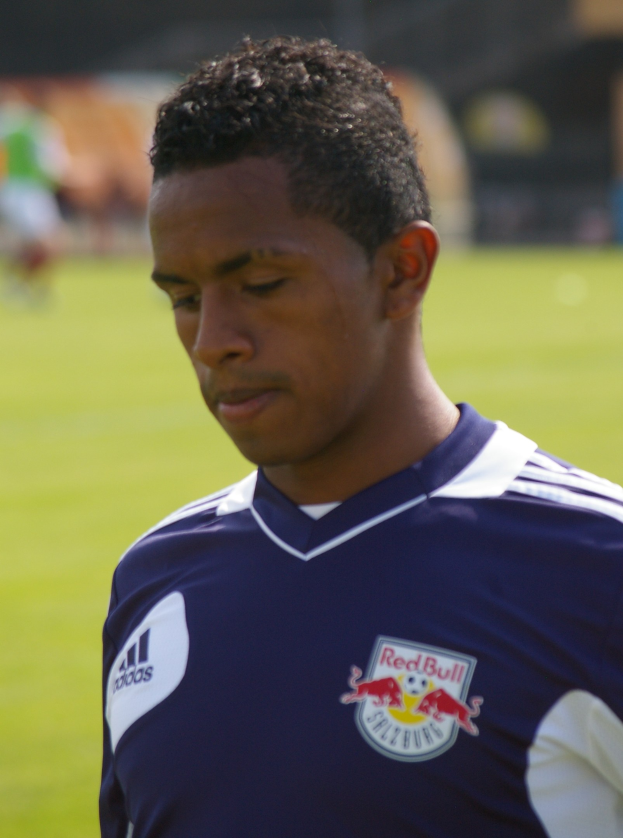 Ein junger Mann in einem blauen T-Shirt steht auf einem Fußballfeld, mit kurzen dunklen Haaren und einem entschlossenen Gesichtsausdruck, der als Flügelspieler mit einem unscharfen Hintergrund erscheint.