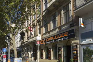 Eine belebte Stadtstraße mit verschiedenen Gebäuden, Fahrzeugen, Fußgängern und Schildern, mit einem hohen Gebäude in der Mitte, das ein "CC Deluxe Cafe & Cocktail Bar"-Schild trägt.
