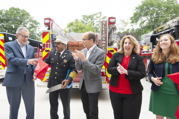 Gruppe von Menschen bei einer Ehrung für die Alexandria Fire Department, klatschend und lachend vor einem Feuerwehrauto mit Bäumen und einem klaren Himmel im Hintergrund.