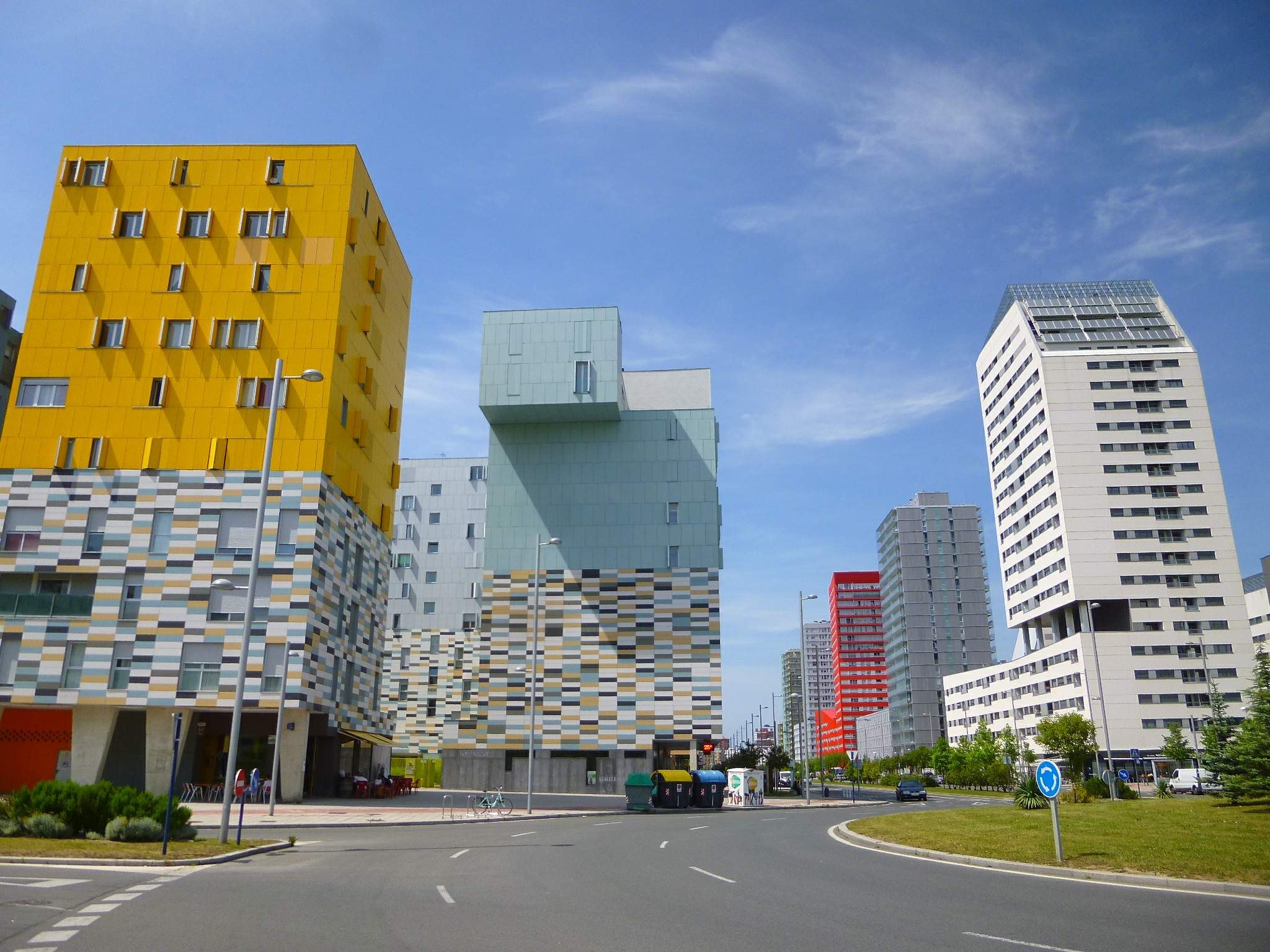 Eine Stadtstraße mit hohen Gebäuden im Hintergrund, Straßenlaternen, Verkehrsschildern, Autos, Büschen, Bäumen und einem bewölkten Himmel, mit den einzigartig gestalteten Gebäuden der Universität Rotterdam.
