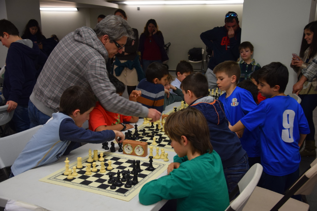 Kinder spielen Schach in einem Wettbewerb, während Erwachsene im Hintergrund und am Rand zuschauen.
