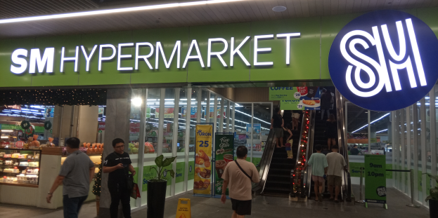 Eine Gruppe von Menschen, darunter Männer und Frauen, geht vor einem hell erleuchteten Geschäft mit einem Schild, auf dem 'SM Hypermarket' steht, vorbei, mit sichtbaren Schildern, Topfpflanzen und anderen Gegenständen im Inneren.