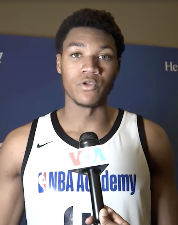 Ein Mann in einem wei├čem Tanktop h├Ąlt ein Mikrofon und spricht mit der Presse bei der NBA All-Star Game 2019, mit einem Banner und einer Wand im Hintergrund.