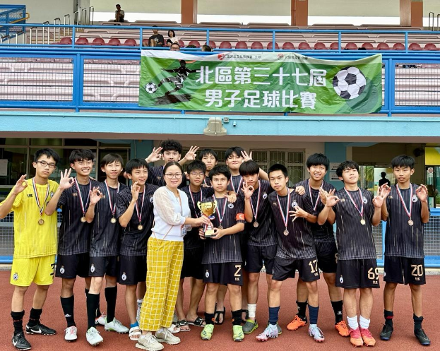 Gruppe junger Männer in Fußballtrikots auf einem Feld, mit Medaillen und einem Pokal, mit einem Banner für die "Yokohama U-16 Jungenfußballmannschaft" im Hintergrund und einigen sitzenden Zuschauern.