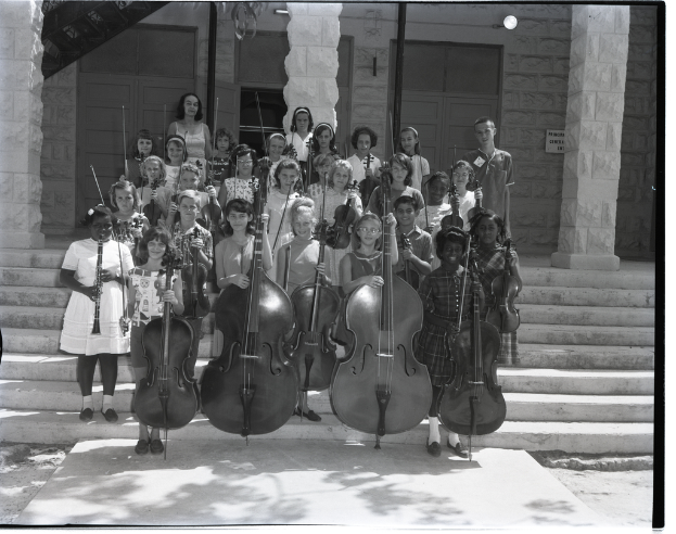 Eine Gruppe von Kindern, die auf einer Treppe stehen und verschiedene Musikinstrumente halten, darunter Celli, Geigen und Trompeten, mit Säulen dahinter und einer Wand mit Türen im Hintergrund.