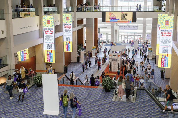 Eine große Menschenmenge, die durch ein großes Gebäude mit Bannern, Topfpflanzen, Rolltreppen, Geländern, Säulen, Texttafeln und Glastüren geht, einige tragen Taschen, wahrscheinlich nehmen sie am Nationalen Buchfestival teil.