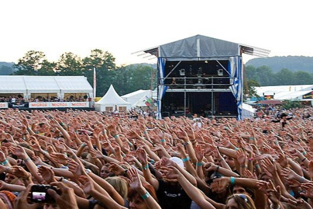 Eine große Menschenmenge auf einem Musikfestival mit erhobenen Händen, einige halten Mobiltelefone, mit einer Bühne, weißen Zelten und Bäumen im Hintergrund bei klarem blauem Himmel.