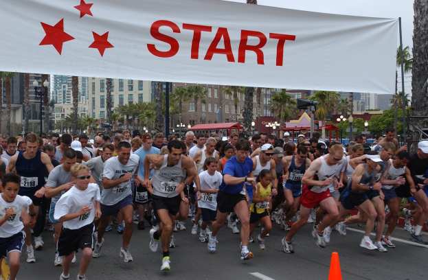 Gruppe von Menschen beim Laufen eines Marathons mit einem Verkehrskegel im Vordergrund und einem Banner mit Text im Hintergrund, umgeben von Bäumen, Laternenmasten, Gebäuden und einem klaren blauen Himmel.