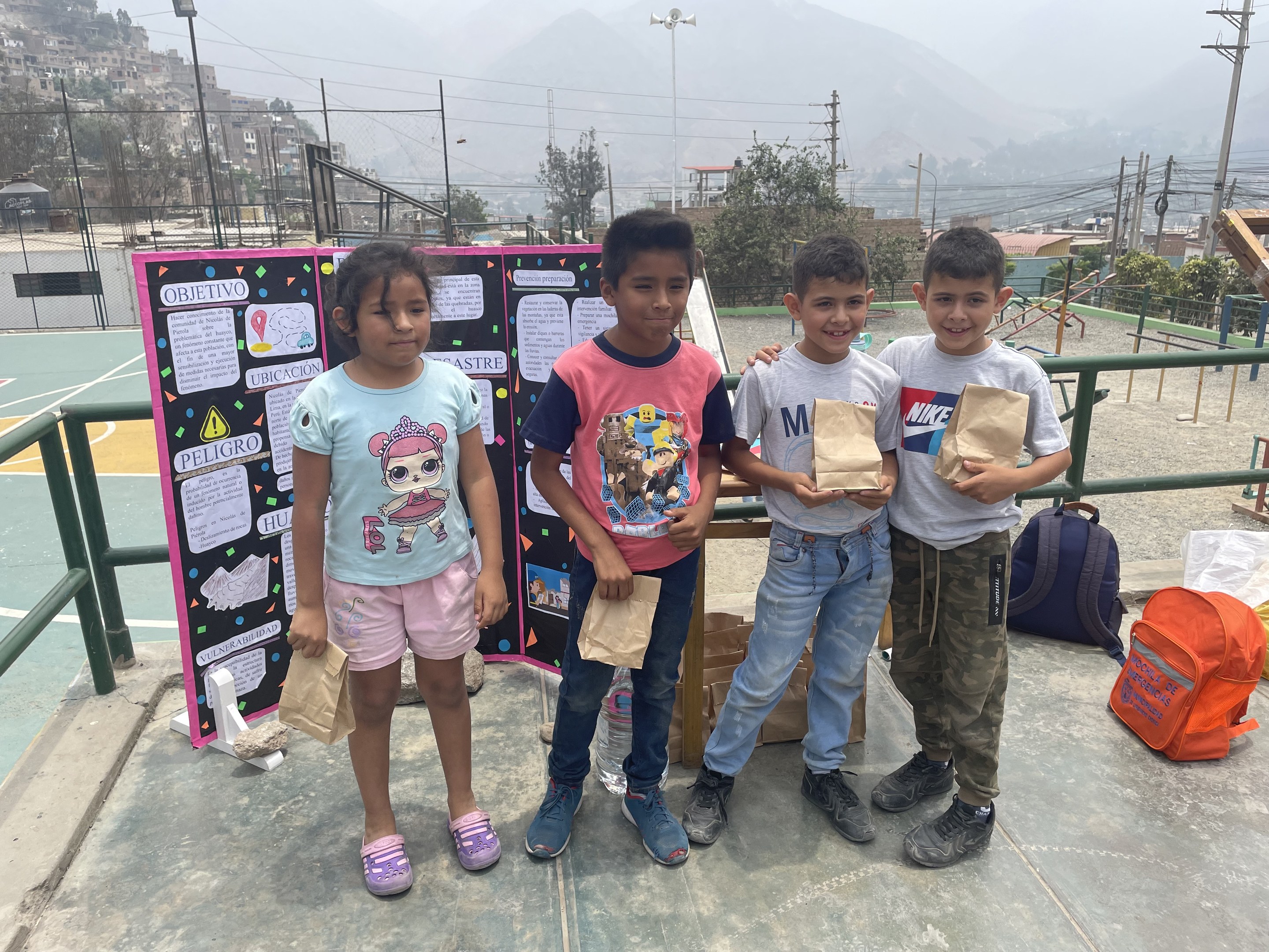 Gruppe von Kindern auf einem Basketballfeld, die Papiertüten halten und links ein Schild mit Text sowie rechts weitere Tüten und Gegenstände auf dem Boden stehen, vor dem Hintergrund von Zäunen, Pfosten, Laternen, Bäumen, Gebäuden, Hügeln und Himmel.