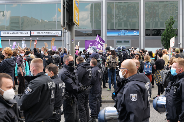 Große Menschenmenge vor einem Gebäude protestierend, einige halten Schilder und tragen Helme, mit einem Schildständer und einem Baum im Vorder- und Hintergrund.