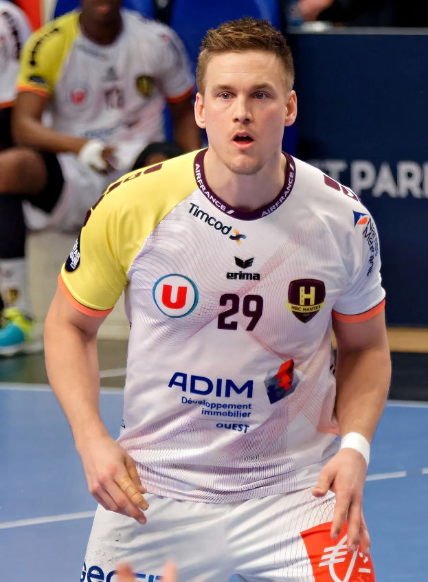 Ein Mann in einem weißen Hemd und gelben Shorts spielt Handball, hält ein Objekt und hat einen entschlossenen Gesichtsausdruck, vor einer Tafel mit der Aufschrift 'Ligue 1 - Futsal - Olympique de Marseille' und einigen Menschen, die auf dem Boden sitzen.