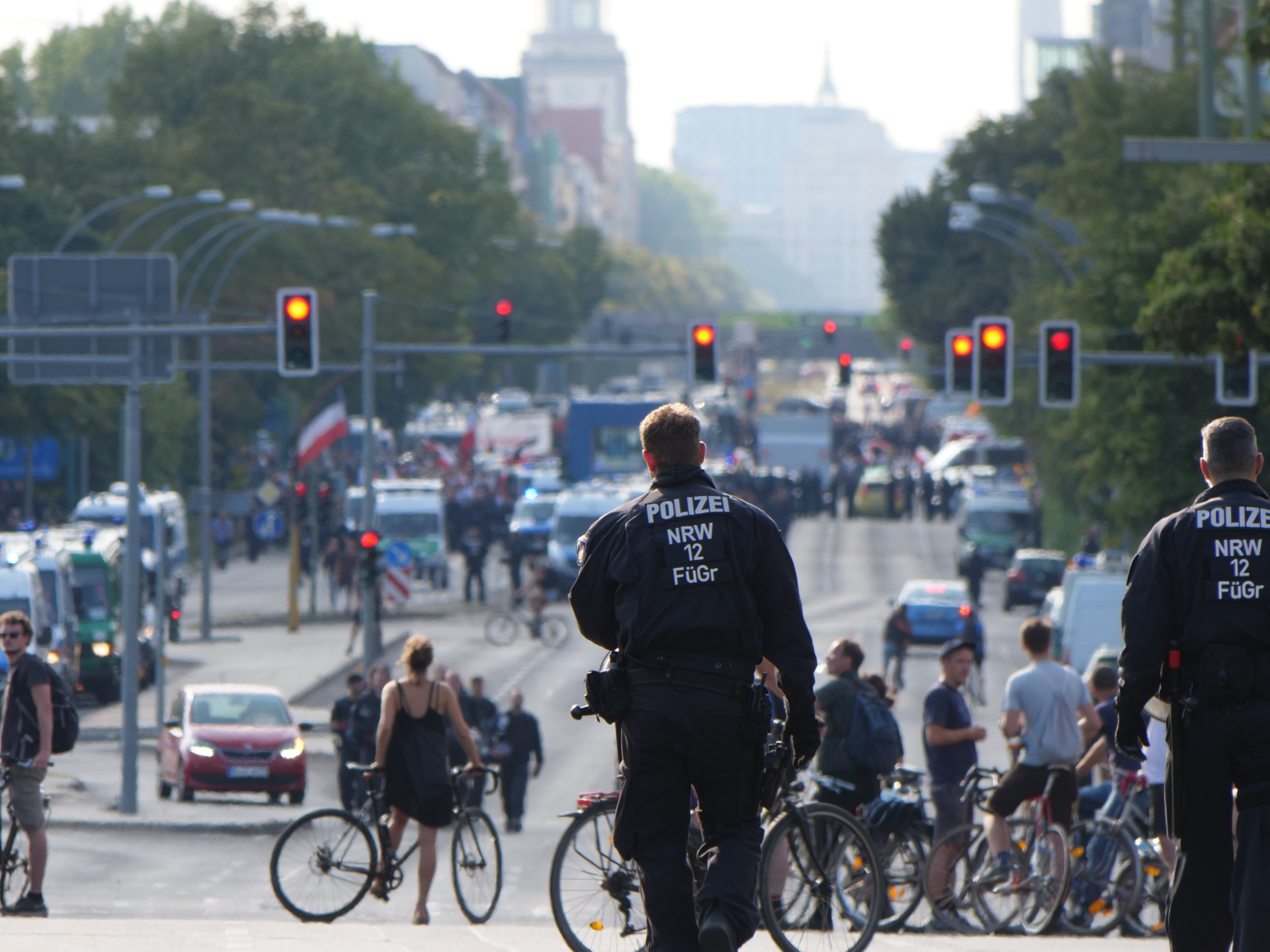 Zwei Polizeibeamte stehen an der Seite einer Straße neben einer Gruppe von Radfahrern, mit Fahrzeugen, Verkehrszeichen, Bäumen, Gebäuden und einem klaren blauen Himmel im Hintergrund.