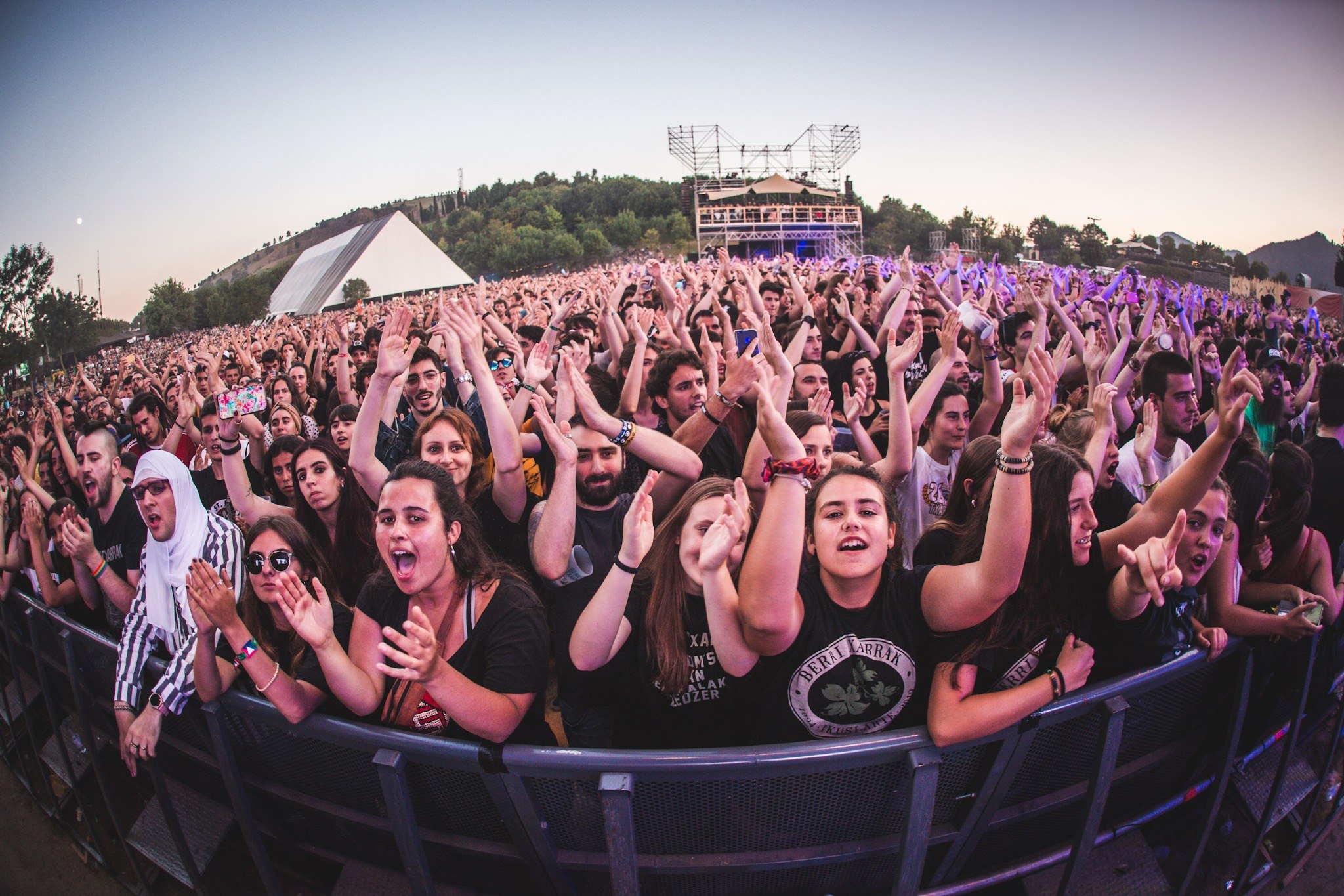 Eine gro├če Menge auf einem Musikfestival mit erhobenen H├Ąnden vor einem Gel├Ąnder, B├Ąumen, Geb├Ąuden und einem klaren blauen Himmel im Hintergrund.