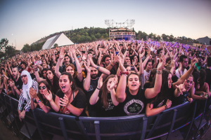 Eine gro├če Menge auf einem Musikfestival mit erhobenen H├Ąnden vor einem Gel├Ąnder, B├Ąumen, Geb├Ąuden und einem klaren blauen Himmel im Hintergrund.