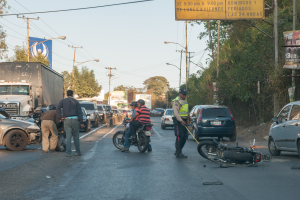 Eine Gruppe von Menschen steht um ein verunglücktes Motorrad auf der Seite einer Straße mit mehreren Fahrzeugen, darunter ein Lastwagen, und einer Hintergrund von Bäumen, Pfählen, Lampen und Schildern unter dem Himmel.